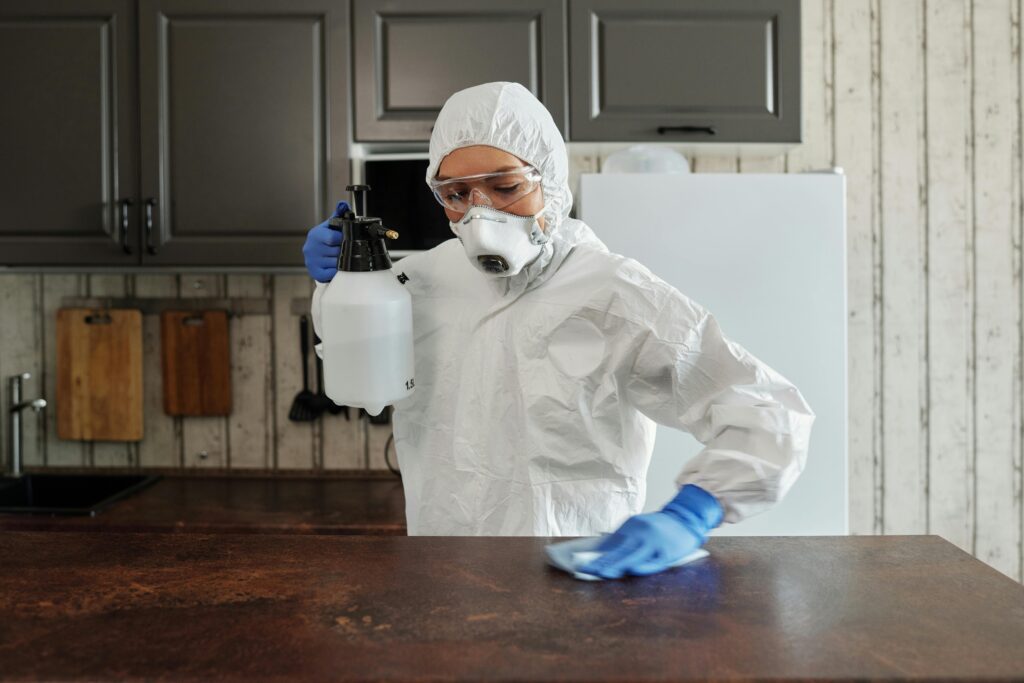 Woman in hazmat suit and mask cleaning table with spray and blue gloves on